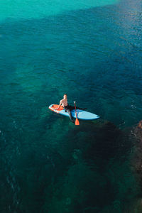 Aerial view of male surfer sitting on paddleboard and floating over rippling azure seawater near rough rocky cliff in sunny day looking at camera