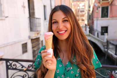 Portrait of smiling young woman holding ice cream against buildings