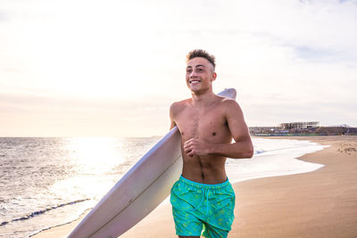 Teenager holding surfboard running on beach against sky