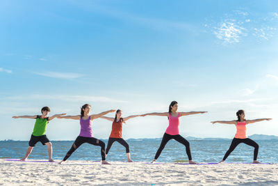 Young friends practicing yoga in warrior position on shore at beach during sunny day