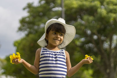 Girl wearing hat holding flowers against tree