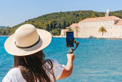 Rear view of young woman using gimbal to take photos and videos on summer vacations by sea