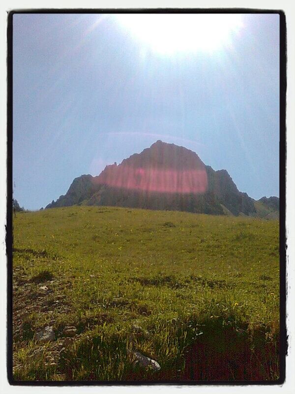 mountain, grass, landscape, sun, sky, tranquil scene, transfer print, tranquility, sunlight, scenics, sunbeam, beauty in nature, built structure, nature, lens flare, mountain range, field, architecture, building exterior, auto post production filter