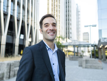 Cheerful elegant adult male manager in formal suit standing near modern building on city street and looking at camera