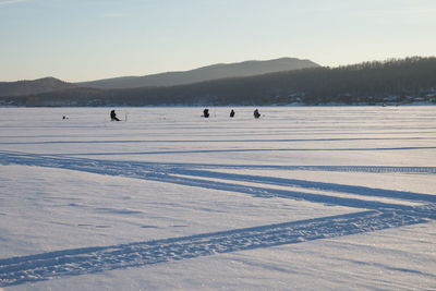 Fishermen sit and fish on a snowy lake, in the evening light.