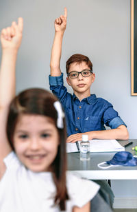 Students with mask on table raising their hands at school. selective focus on boy in background