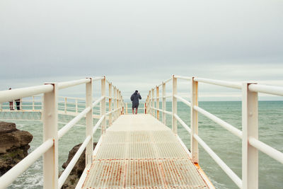 Rear view of man walking on pier over sea against cloudy sky