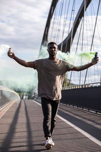 Full length of man holding distress flare walking on footbridge against sky