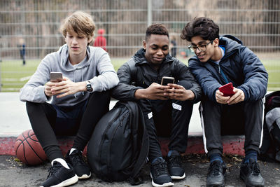 Social media addicted friends using mobile phones while sitting on sidewalk after basketball practice in city