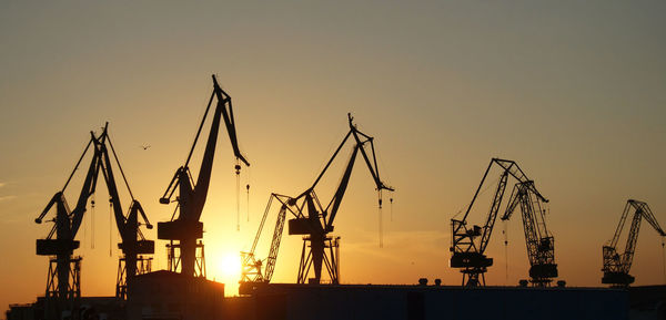 Silhouette cranes at commercial dock against sky during sunset