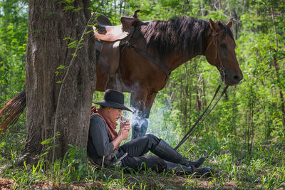 Side view of cowboy smoking cigarette while sitting by horse at forest