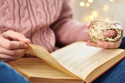 Midsection of woman holding coffee cup reading book at home