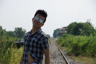 Portrait of handsome man wearing sunglasses while standing on railroad tracks against sky