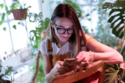 Young successful female plant shop owner using smartphone, responding customer in social media