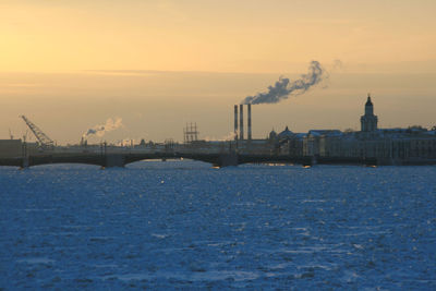 Smoke emitting from factory by river against sky during sunset