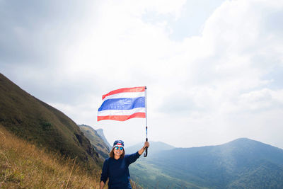 Happy woman waving thai flag on mountain against cloudy sky