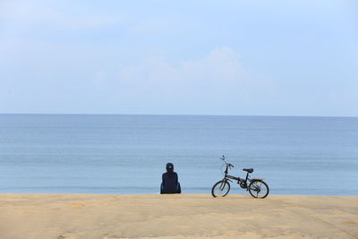 Woman with bicycle sitting at beach against clear sky