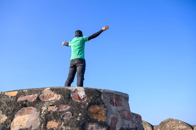 Low angle view of man standing on rock against clear blue sky