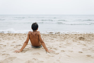 Rear view of shirtless young man relaxing at beach against clear sky