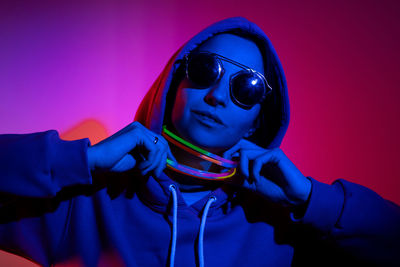 Cyberpunk woman in a hooded hoodie and sunglasses dances against a wall with neon sticks hanging 