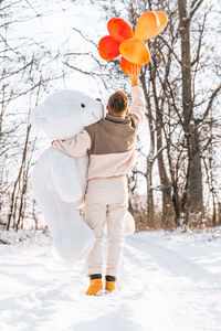 Rear view of man holding stuffed toy with balloon standing on snow covered land