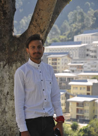 A college student looking at camera standing against tree with college campus in background 