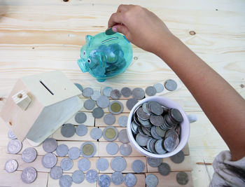 Person holding coin over piggy bank by wooden model home and cup on table