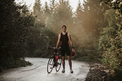 Full length of young man with bicycle walking on road against plants