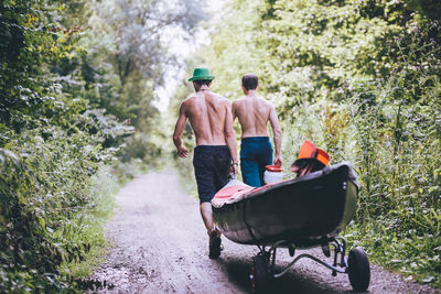 Rear view of shirtless friends carrying boat in forest