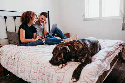 Young couple using laptop while dog sleeping on bed at home