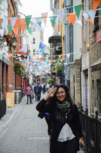 Portrait of smiling young woman saluting while standing on street amidst buildings