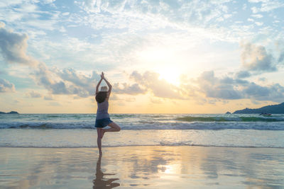 Full length rear view of woman doing tree pose at beach against sky during sunset