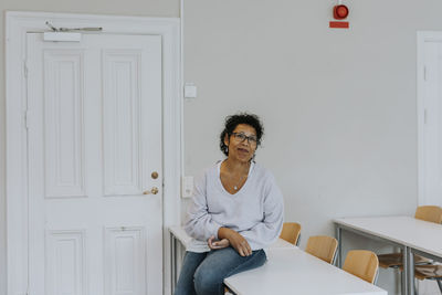 Smiling mature teacher sitting on desk in classroom