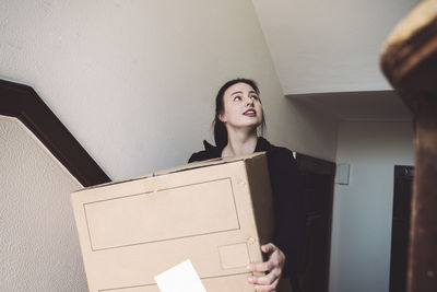 Confident delivery woman with cardboard box looking up against wall