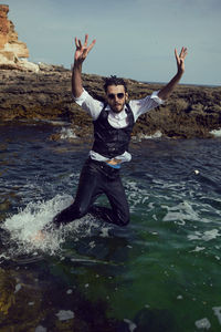 Man with a beard and sunglasses in clothes a vest and a white shirt jump in the sea among the rock