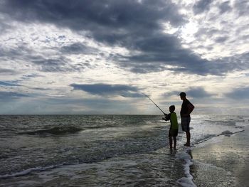 Father and boy fishing on shore at beach against sky
