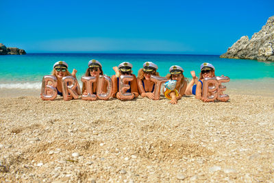 Portrait of smiling bride with bridesmaid lying at beach against clear sky