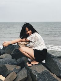 Young woman sitting on rock by sea against clear sky