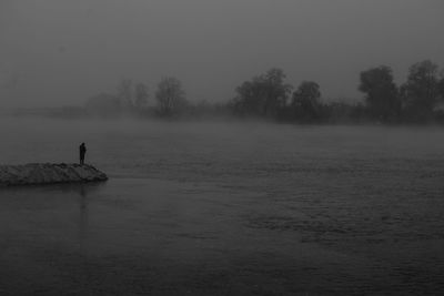 Silhouette person standing on rock at lake at dusk during foggy weather