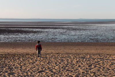 Rear view of boy standing on sand at beach against sky