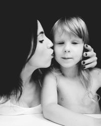 Mother kissing shirtless daughter against black background