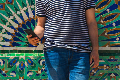 Midsection of man using mobile phone while standing against patterned wall 