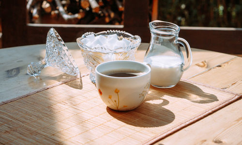 A cup of coffee stands outside on a table next to a jug of milk and a sugar bowl on a sunny day