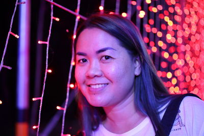 Close-up portrait of smiling woman by illuminated lighting equipment at night