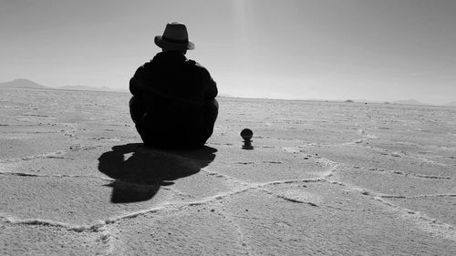 Rear view of silhouette man sitting on sand at beach against sky