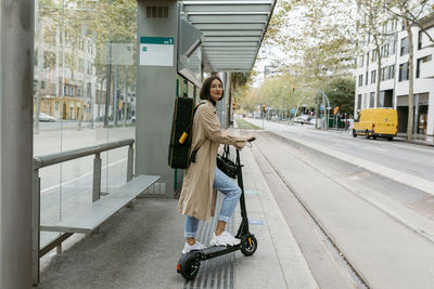 Woman with push scooter and instrument case standing at tramway station in city