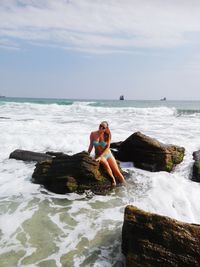 Full length of woman in bikini sitting on rock amidst sea against sky