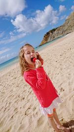 Girl eating strawberry while standing at beach against sky during sunny day