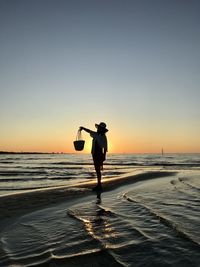 Woman holding purse while standing at beach against sky during sunset