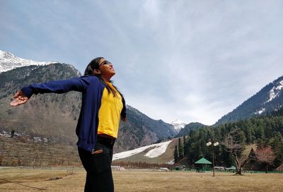 Smiling carefree woman with arms outstretched standing against mountains